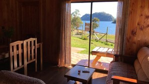 A wooden cabin with a large sliding glass door leading to a deck. Inside, there is a wooden chair, a small table with a remote control, and a sofa. - beach shore hut (lago ranco)