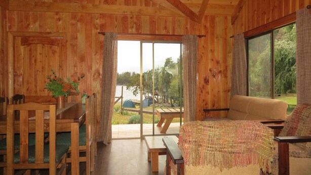 A wooden cabin interior with a sofa, a dining table, and chairs. There is a view of a lake and trees through the glass doors. - beach shore hut (lago ranco)