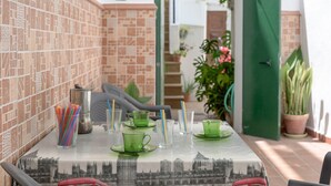 Ein Esstisch mit grünem Glas und einer Tischdecke mit Stadtansicht, umgeben von Blumentöpfen und einer grünen Tür.