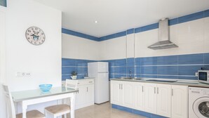 Une cuisine moderne avec des meubles blancs, un crédence en carreaux bleus, un four à micro-ondes, un lave-linge et un coin repas avec une table en verre et deux chaises.