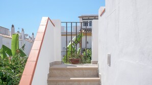 Un escalier menant à un balcon orné d’un pot de fleurs, avec une rambarde en métal et un mur blanc.