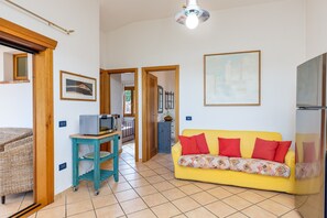 Un salon avec un canapé jaune, un fauteuil en osier, une petite table sur laquelle se trouve un micro-ondes, et une cuisine en arrière-plan.