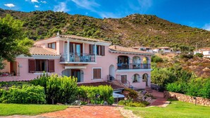 Una villa rosa con un camino de piedra, un balcón y montañas al fondo.