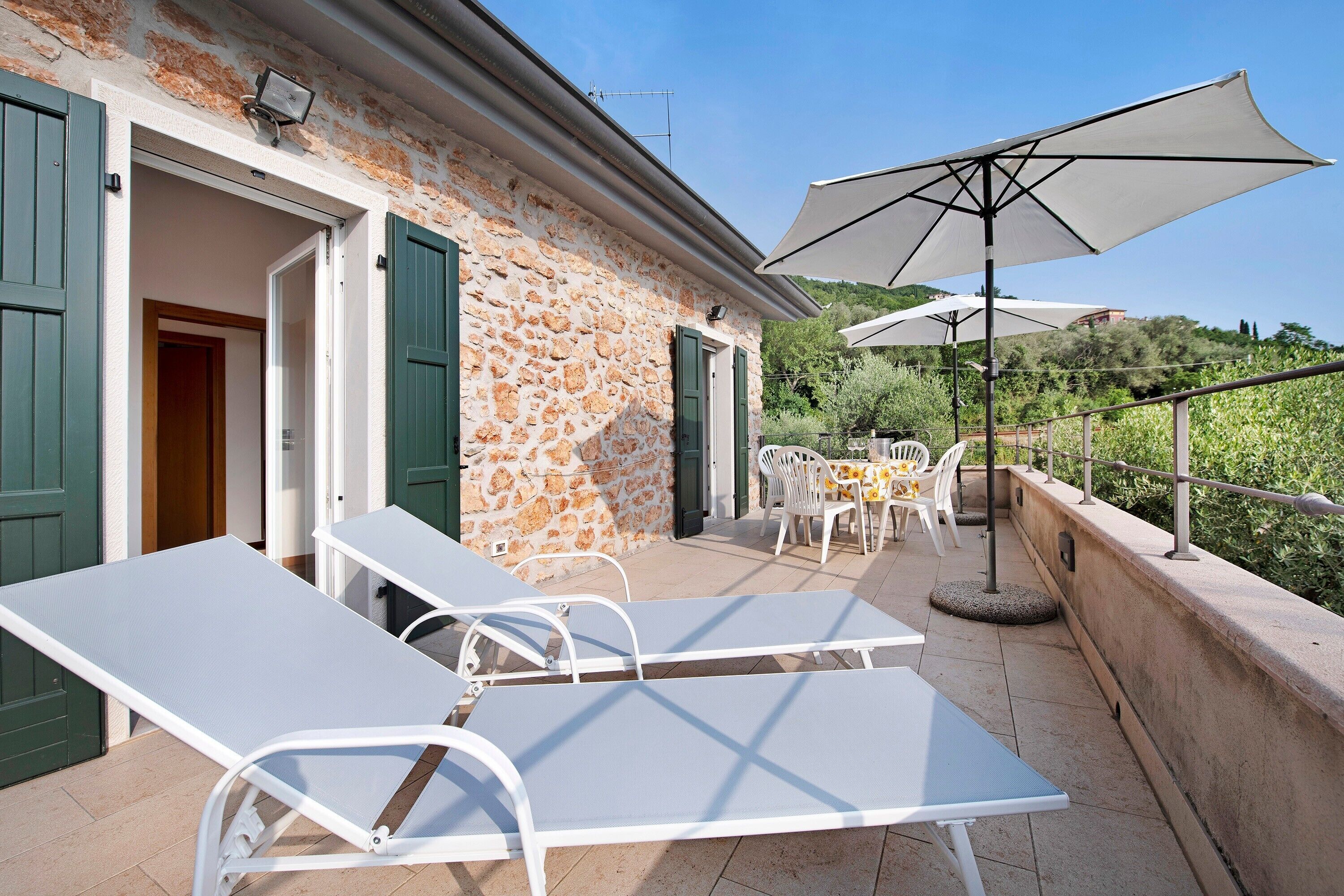 Eine Terrasse im Freien mit Lounge-Möbeln und Sonnenschirmen, ein Steinhaus mit grünen Fensterläden und Blick auf viel Grün.
