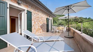 Eine Terrasse im Freien mit Lounge-Möbeln und Sonnenschirmen, ein Steinhaus mit grünen Fensterläden und Blick auf viel Grün.
