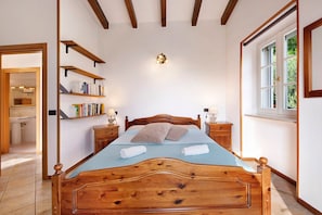 Un letto in legno con testiera, un comodino con lampada, uno scaffale con libri e una finestra vista verde.