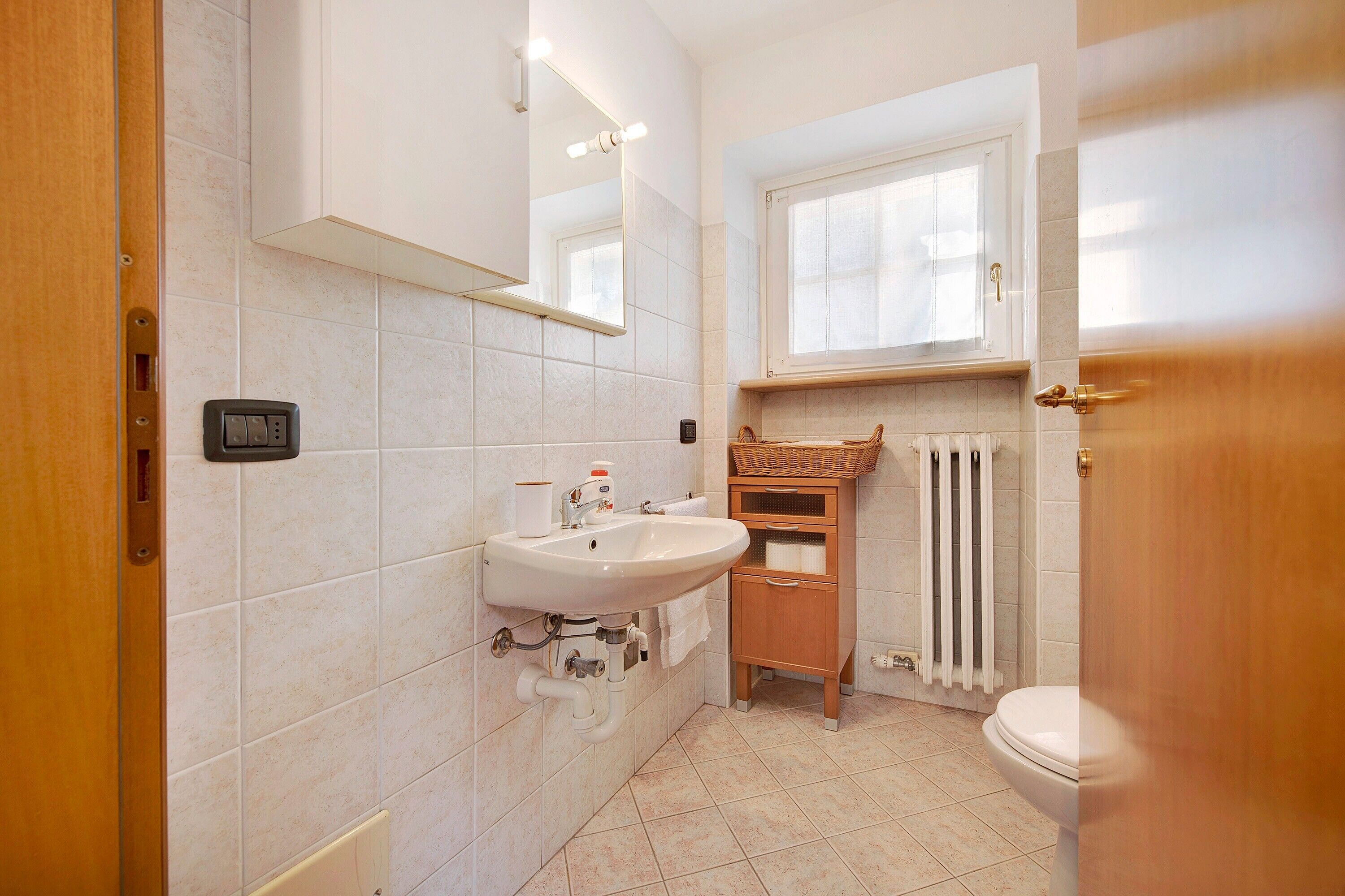 A bathroom with a sink, mirror, window, radiator, and wooden door.