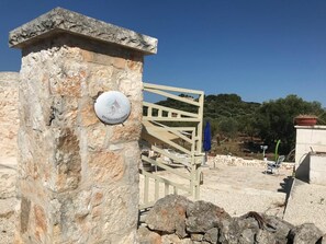Um pilar de pedra com uma placa, um portão de madeira e uma área de praia com guarda-sóis e espreguiçadeiras.
