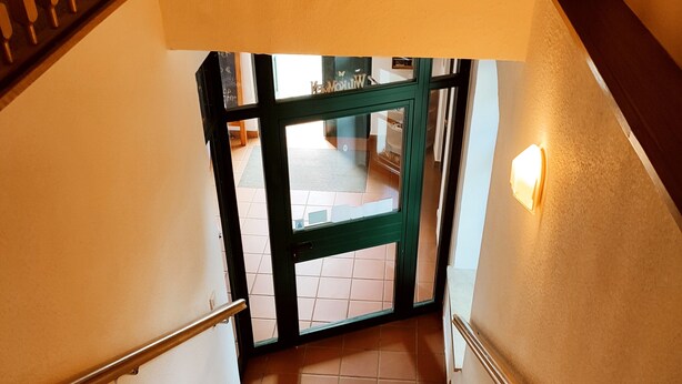 Una escalera con barandilla que conduce a un espacio abierto con suelo de baldosas y una puerta verde.