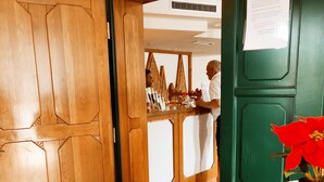 Habitación de hotel con una puerta de madera, una puerta verde, piso de baldosas y una mesa blanca con una flor roja.