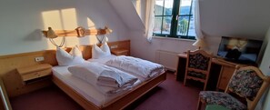 Una cama bien hecha con sábanas blancas, cabecera de madera y mesita de noche con lámpara. Hay una silla con cojín estampado, un escritorio con lámpara y un televisor sobre un mueble de madera.