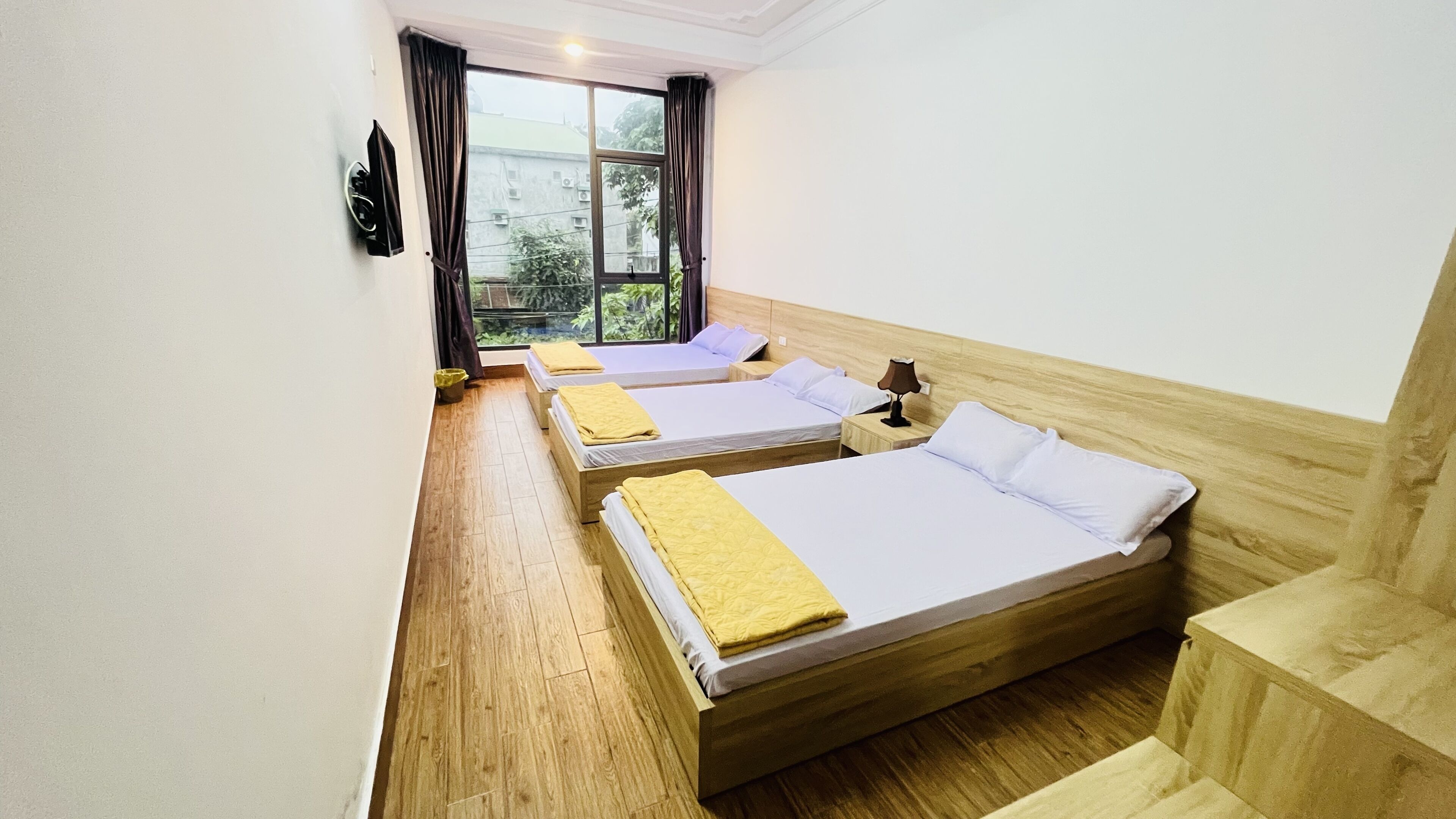 A hotel room with two single beds, wooden flooring, a window with curtains, and a view of greenery outside.