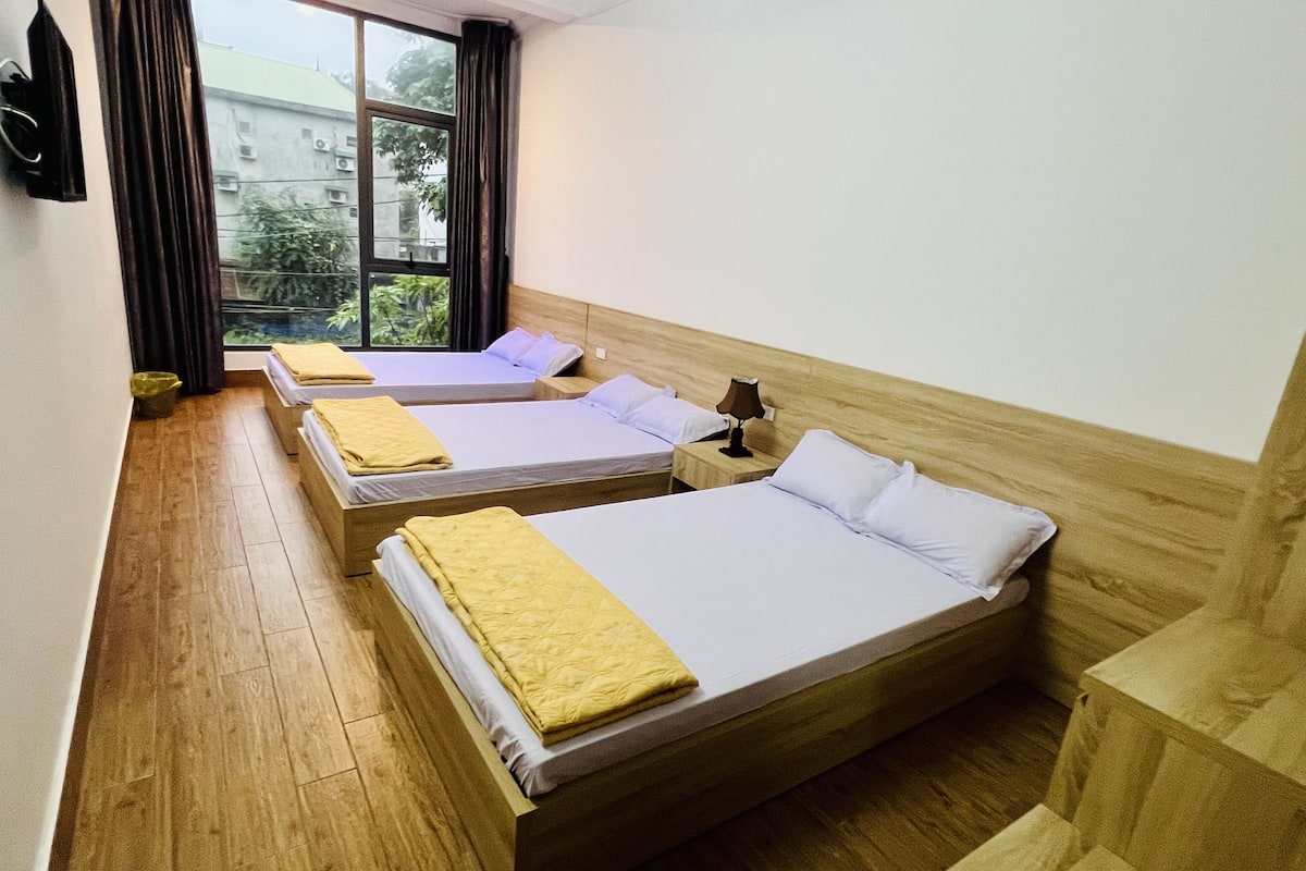 A hotel room with two beds, wooden flooring, a nightstand with a lamp, and a window with curtains.
