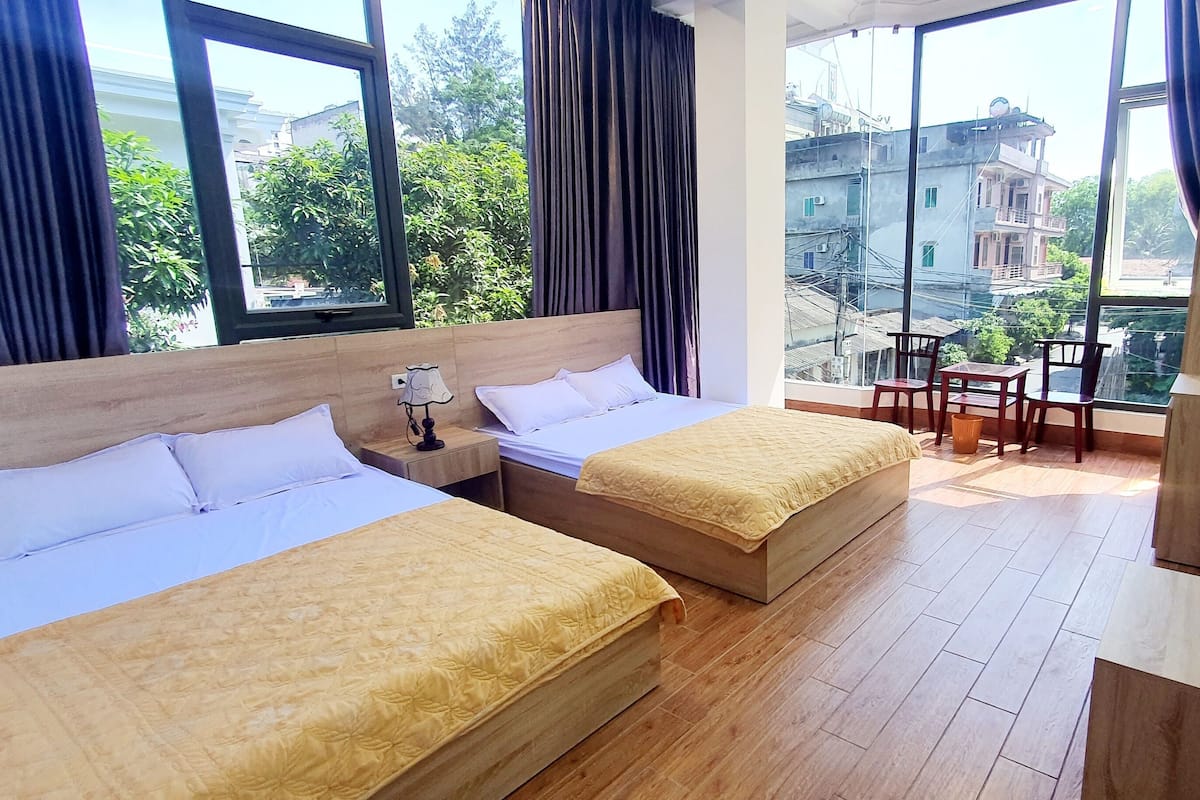 A hotel room with two beds, a small table with chairs, a TV, and a view of buildings and trees.