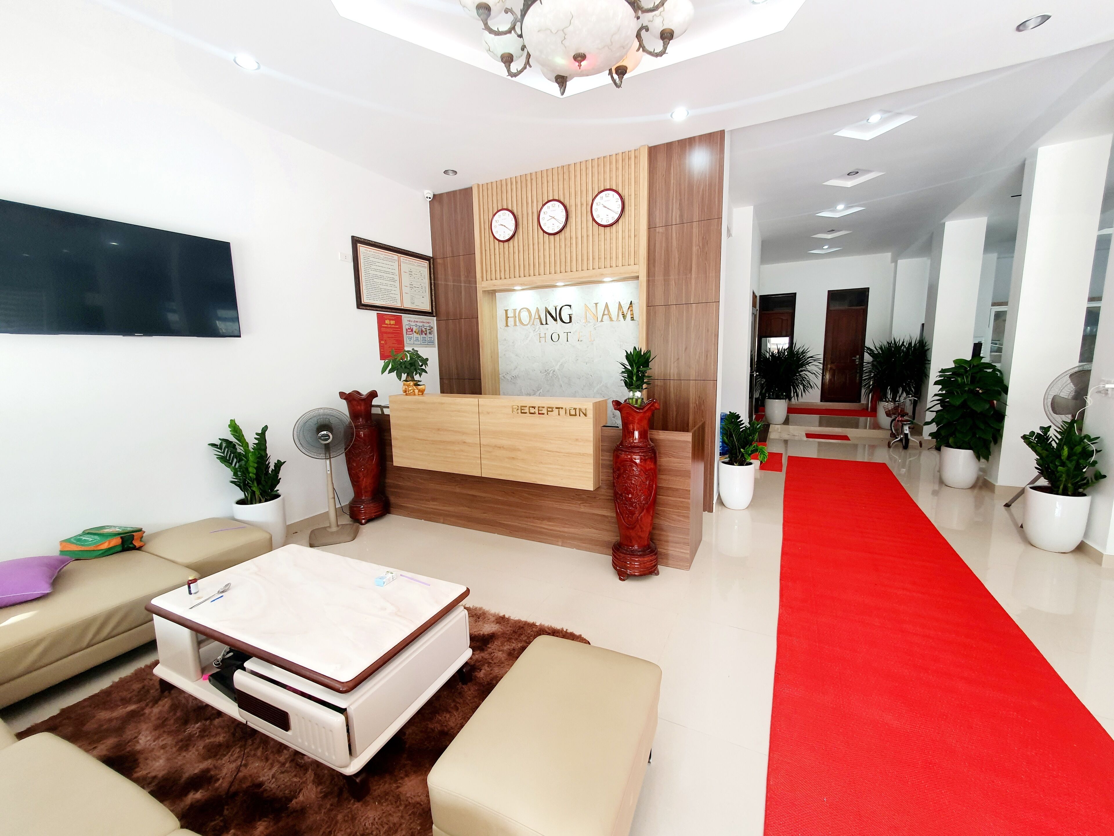 A hotel reception area with a wooden reception desk, a flat-screen TV, and a red carpet leading to the entrance.