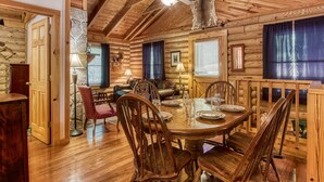 Interior de una cabaña de troncos con comedor, chimenea y ventilador de techo.