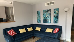 Un salon moderne avec un canapé d’angle bleu foncé, un pouf assorti et trois tableaux abstraits accrochés au mur.