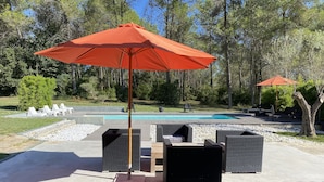 Een terras buiten met een zwembad, omringd door bomen, met rieten stoelen en een oranje parasol.