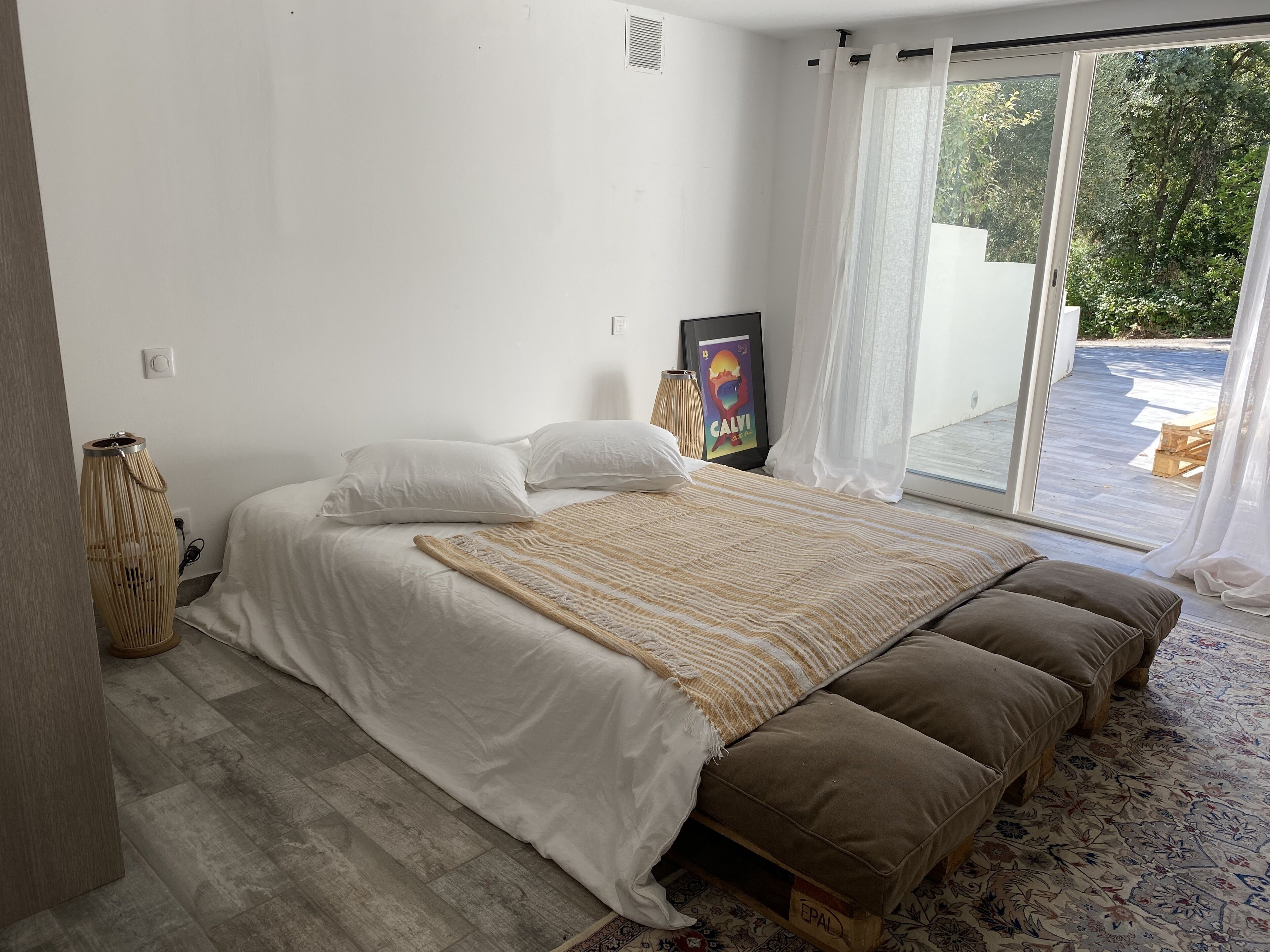 A bedroom with a large bed, a window with sheer curtains, a framed picture on the wall, and a wooden floor.