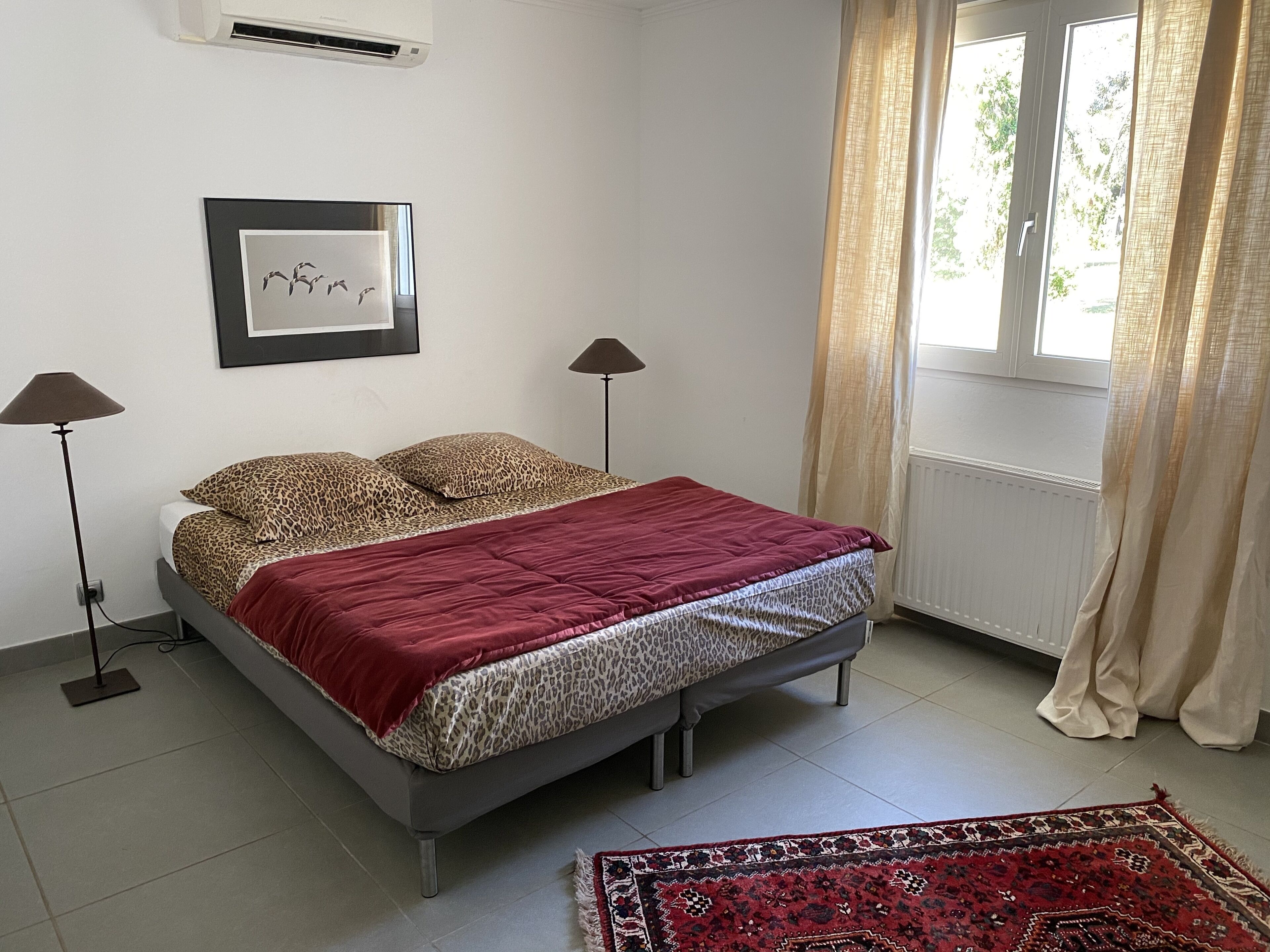 Une chambre à coucher avec un lit, deux lampadaires, un tableau encadré au mur, une fenêtre avec des rideaux et un tapis à motifs sur le sol.