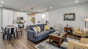 A modern living room with a gray sofa, wooden coffee table, and patterned armchairs. A kitchen with white cabinets and a dining area with a table and chairs is visible.