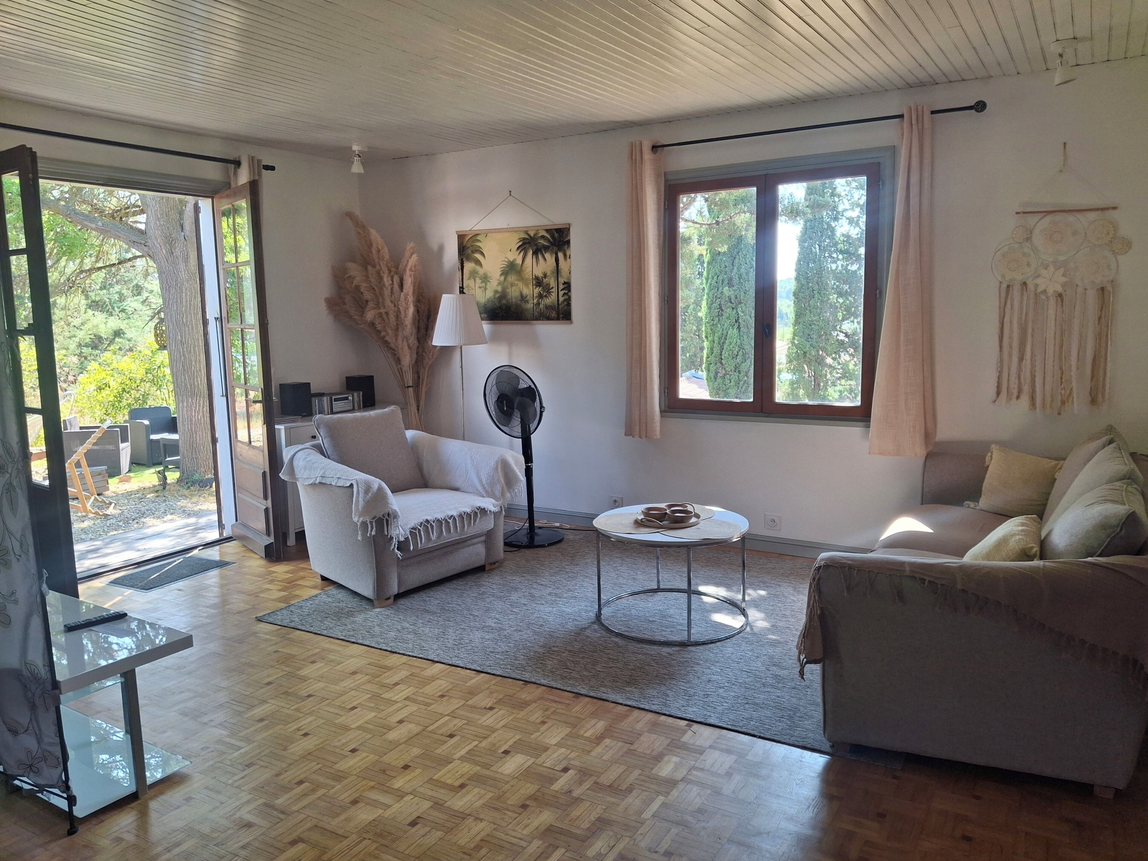 Een woonkamer met een bank, fauteuil en een salontafel. Er is een ventilator, een lamp en een schilderij aan de muur.