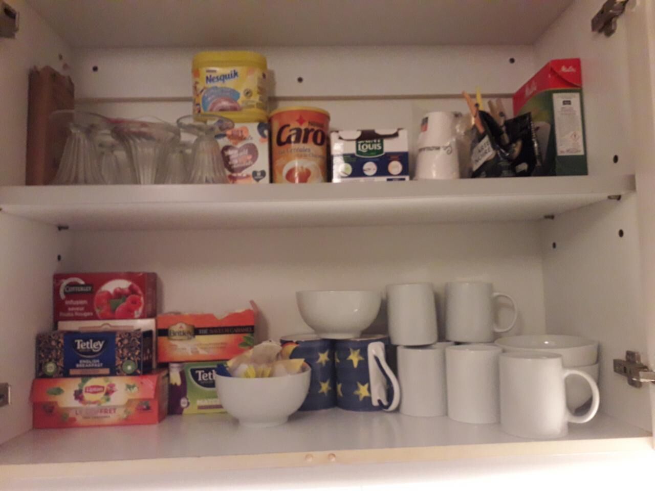 A kitchen pantry with shelves stocked with cereal boxes, a can of baby formula, and white mugs.