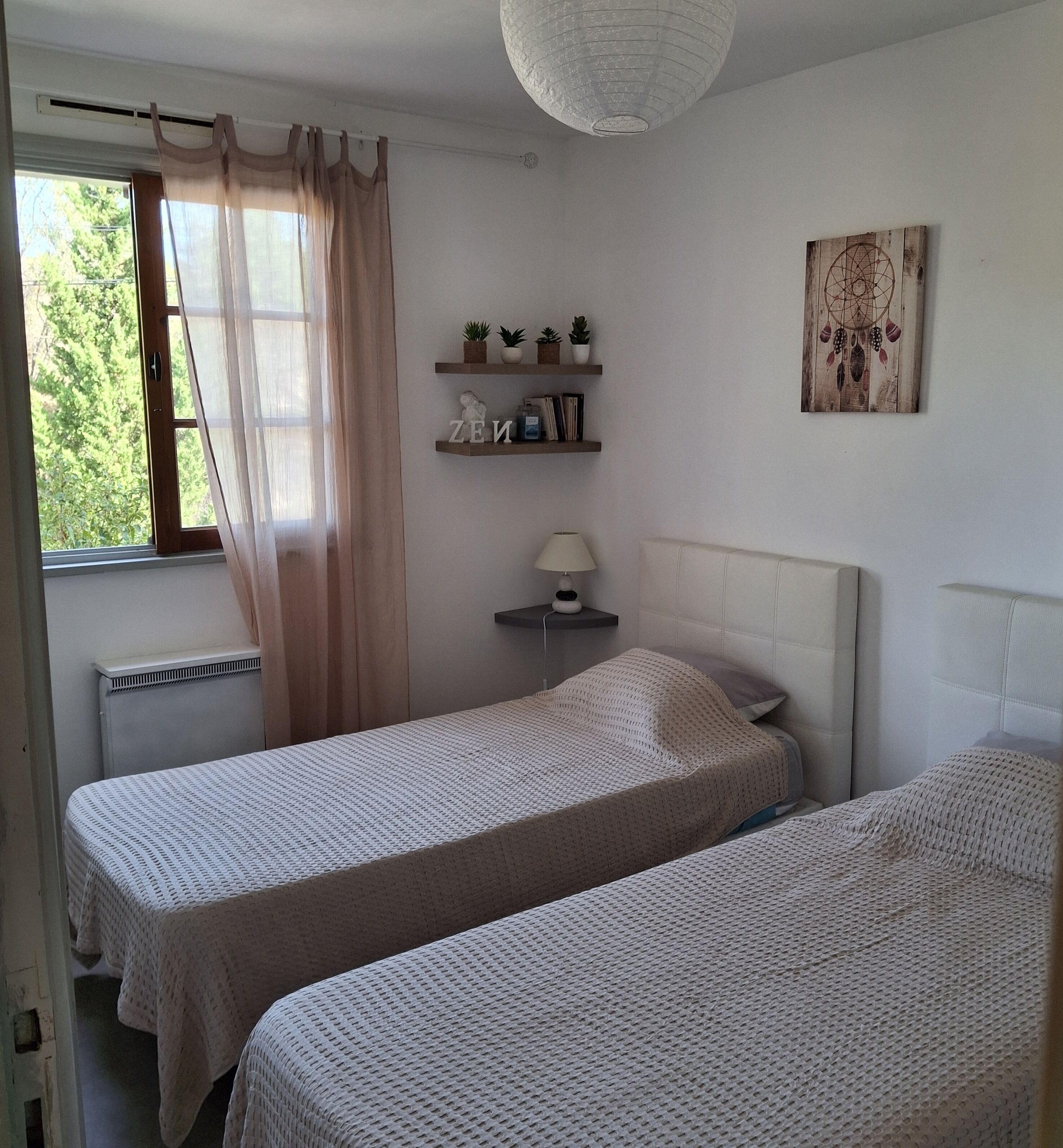 Een slaapkamer met twee eenpersoonsbedden, een nachtkastje met een lamp, een plank met decoratieve objecten en een raam met gordijnen.