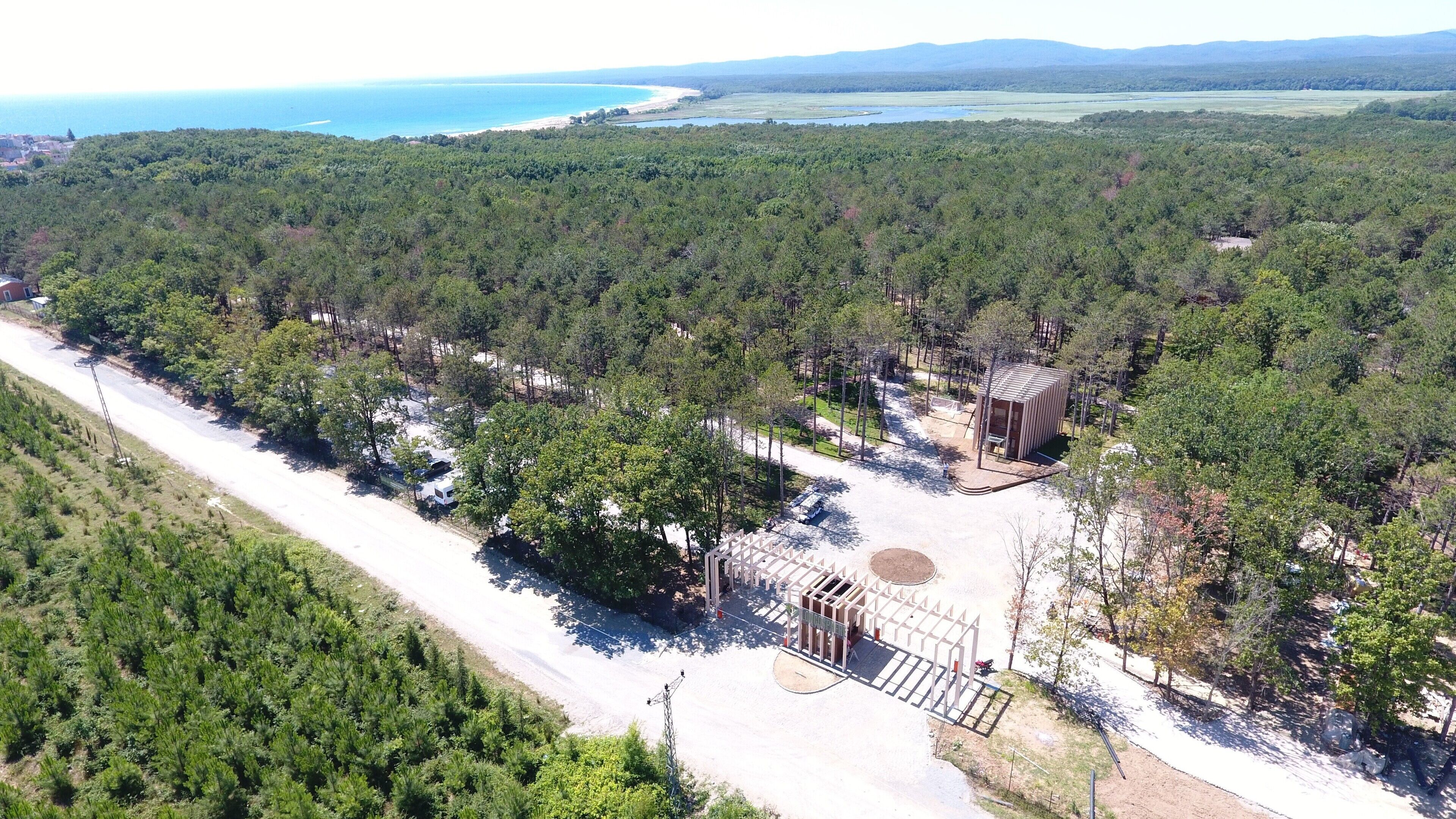 An aerial view of a forested area with a paved road, a small building, and a beach in the background.