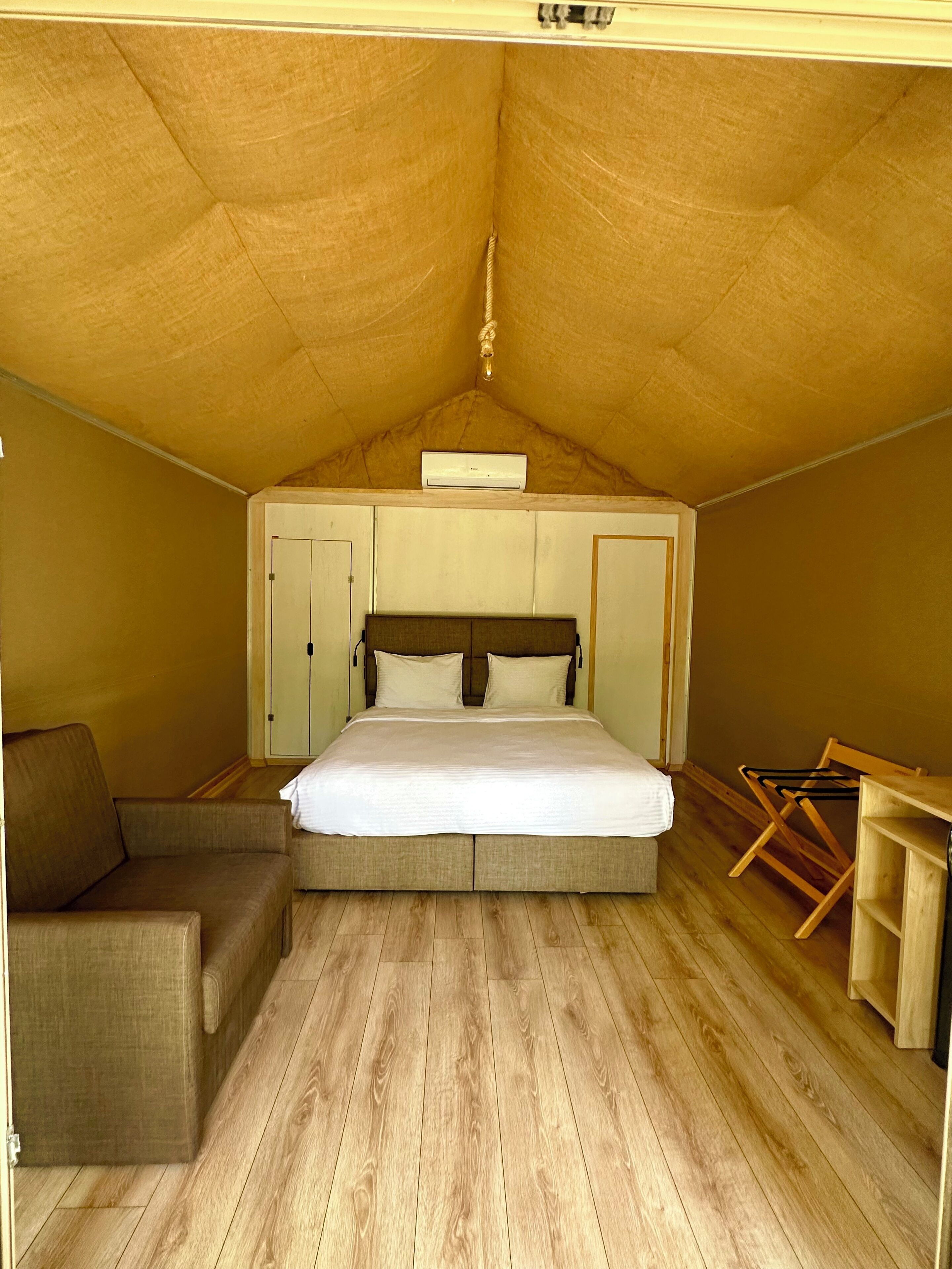 Un dormitorio con techo de madera, una cama con ropa de cama blanca, piso de madera, un pequeño sofá y una silla de madera.