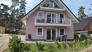 Une maison à deux étages avec un balcon, une terrasse dotée de mobilier d’extérieur et un jardin qui l’entoure.