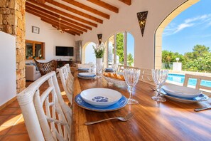 A dining area with a long wooden table set for a meal, a stone wall, and a view of a swimming pool.