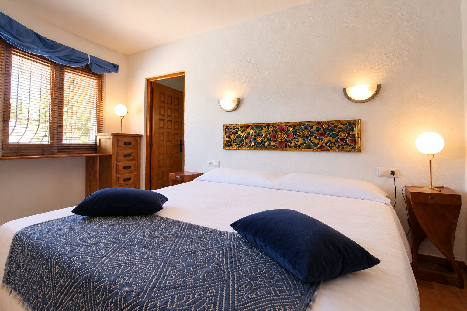 A bedroom with a bed, two bedside lamps, a wooden cabinet, a window with blinds, and a decorative wall piece.