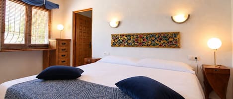 A bedroom with a bed, two bedside lamps, a wooden cabinet, a window with blinds, and a decorative wall piece.
