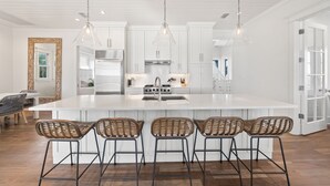 A modern kitchen with white cabinetry, a central island with bar stools, pendant lighting, and a view into the dining area. - Stunning Retreat w/Gulf views, heated pool, beach access, and bikes. Sleeps 12! (Seagrove Beach)