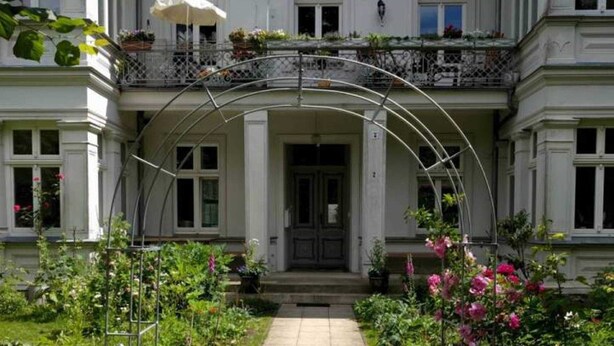 Un edificio clásico con una entrada central, balcones con barandillas y un jardín lleno de flores.