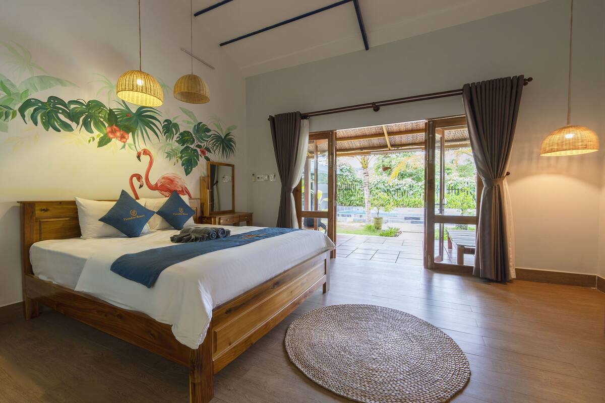 A bedroom with a wooden bed, a large window with curtains, a woven rug, and a wall mural of a flamingo and plants.