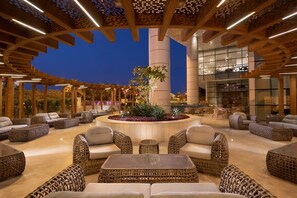 A spacious outdoor lounge area with wicker furniture, a central plant arrangement, and a modern architectural design with wooden beams and glass walls. - Le Méridien Riyadh (Riyadh)