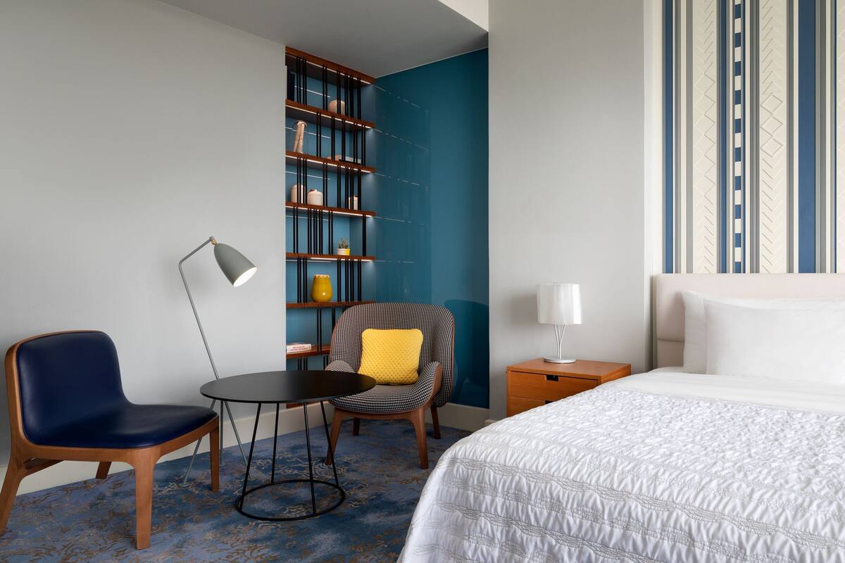 Una habitación de hotel con una cama, un escritorio, una silla, una estantería y una lámpara.