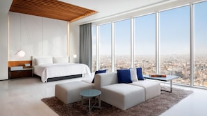 Ett modernt hotellrum med en stor säng, en soffa, ett litet bord och utsikt över staden genom stora fönster.