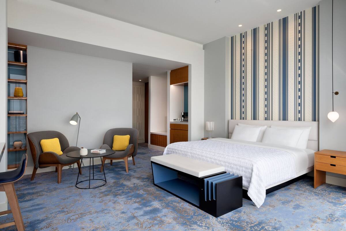 Una habitación de hotel moderna con una cama grande, dos sillones, una mesita y una estantería.