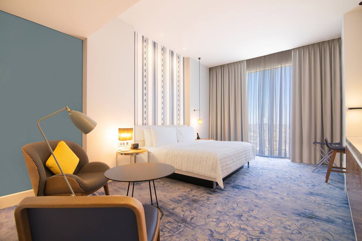 Habitación de hotel con cama, escritorio, silla, lámpara y vistas al exterior.
