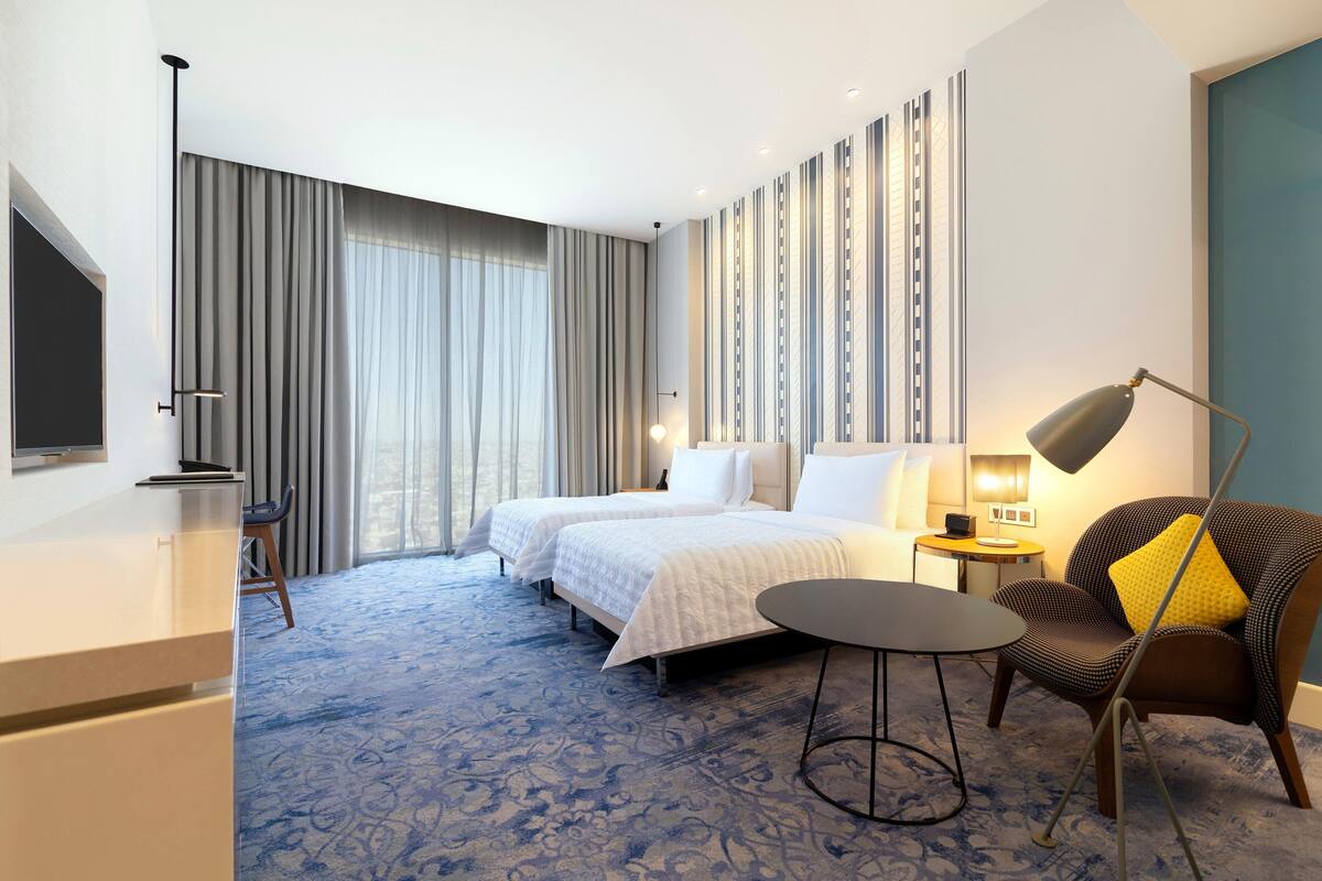 Habitación de hotel con dos camas, un escritorio, una silla, una lámpara y vistas a la ciudad.