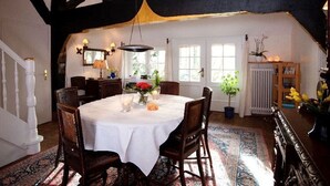 A dining area with a round table, chairs, and a chandelier. There are windows with curtains, a radiator, and a plant. - Semi-detached house FeWo 1 - holiday homes Ohlenbusch (Worpswede)