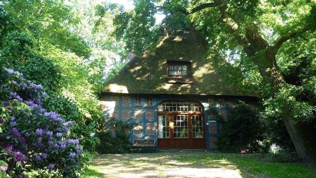 A rustic house with a thatched roof, surrounded by lush greenery and flowering plants. - Semi-detached house FeWo 1 - holiday homes Ohlenbusch (Worpswede)