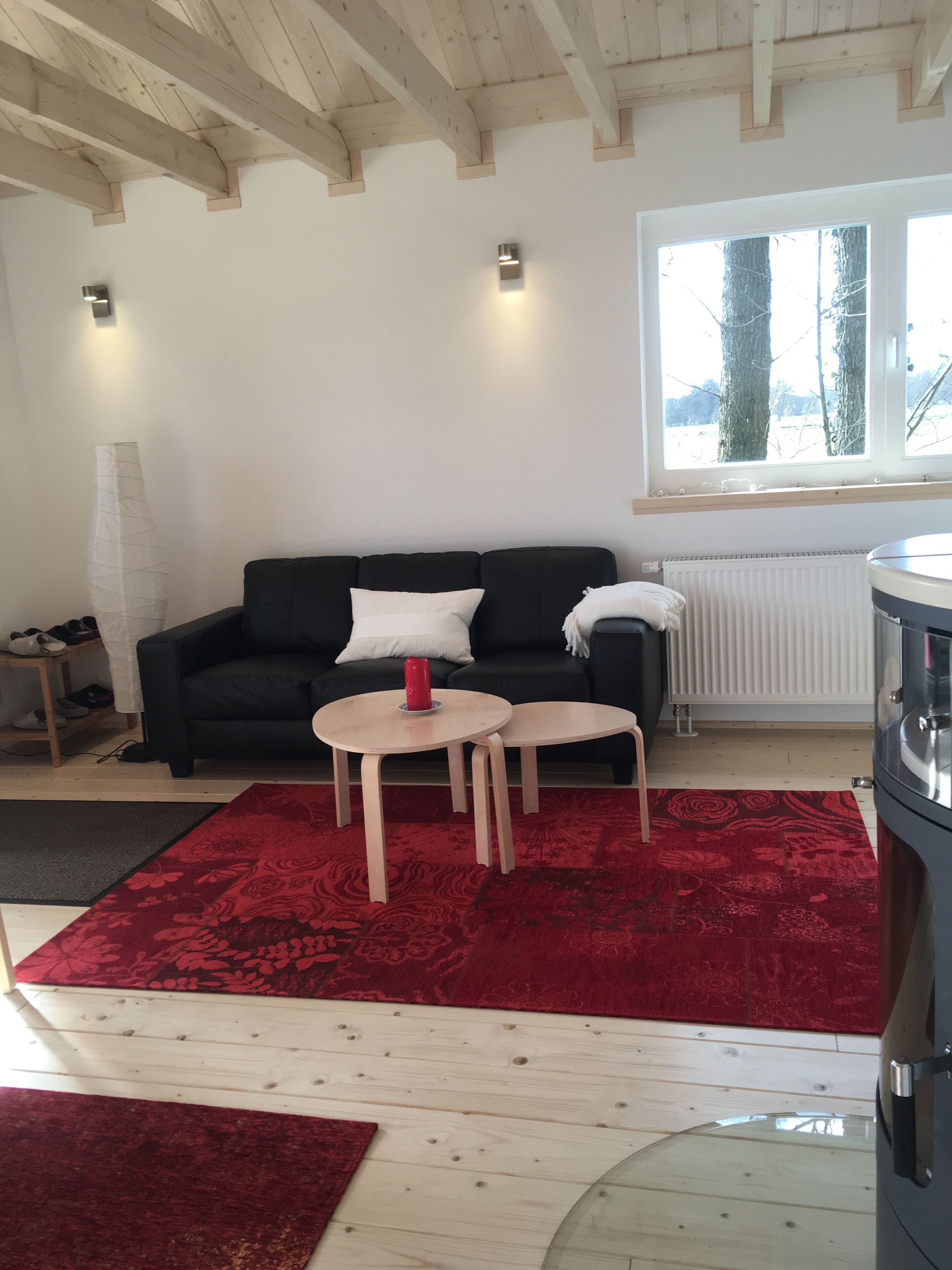 A modern living room with a black sofa, two small wooden tables, a red patterned rug, and a large window with a view of trees.