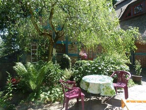 An outdoor patio with a round table and two chairs, surrounded by lush greenery and a brick building with windows.