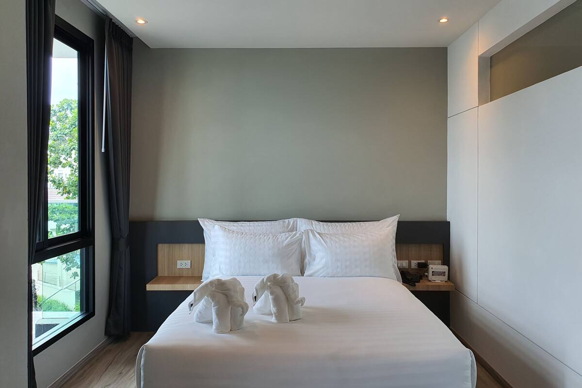 Hotellihuone, jossa on suuri sänky, kaksi tyynyä, yöpöytä, jossa on puhelin ja lamppu, sekä ikkuna, jossa on verhot.