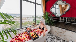 A woman in a bathtub filled with rose petals and fruit, with a view of a cityscape through large windows. - Athena Hotel (Quy Nhon Nam)