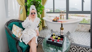 A room with a large bathtub, a green armchair, and a view of the city. - Athena Hotel (Quy Nhon Nam)